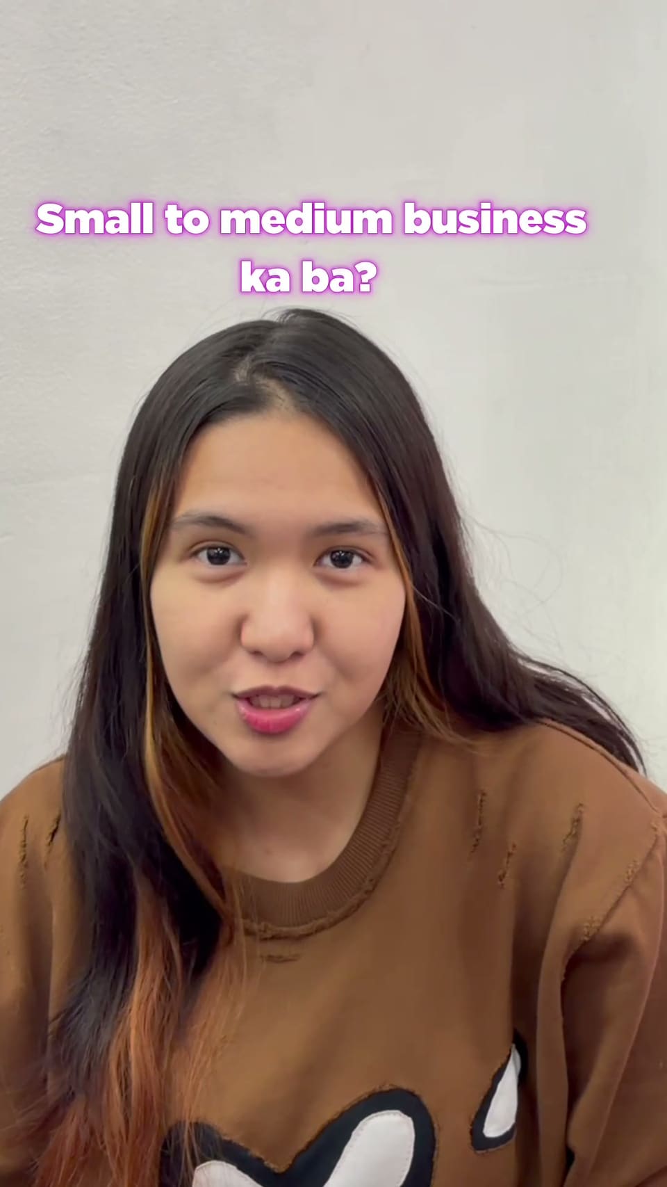 Vertical video frame of a woman in a brown sweater against a white wall, with a pink-outlined caption “Small to medium business ka ba?”.